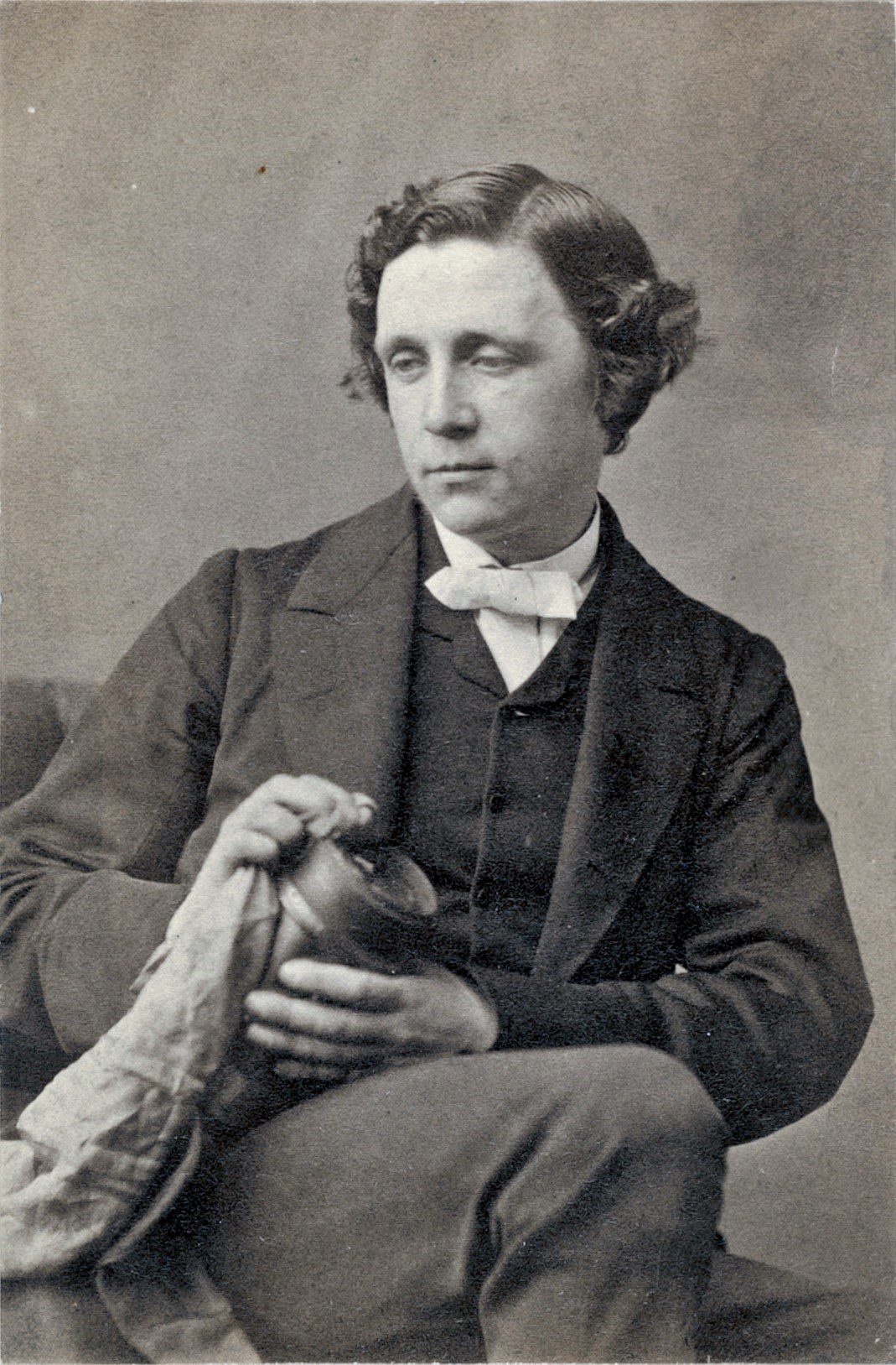 Sepia photograph of Lewis Carroll (Charles Lutwidge Dodgson), seated in a dark jacket and white bow tie, holding a camera lens and looking down to one side, taken by Oscar Rejlander in 1863